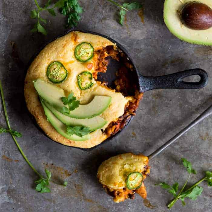Enchilada bake. Small skillet with baked cornbread and chicken enchilada mix topped with thinly sliced jalapenos, avocado slices and cilantro. Single spoonful removed and on the side