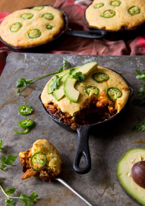 Enchilada bake. small cast iron skillet filled with chicken enchilada mixture and baked with a cornbread and jalapeno topping. Garnished with avocado slices and cilantro. Spoonful removed and is placed in front. 