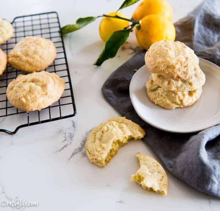 Cooling rack with lemon cookies on it next to a white plate with a stack of 3 lemon cookies on a gray napkin. Whole lemons in the background. A cookie with a bite taken out in the foreground