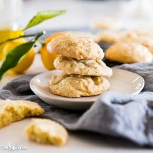 Stack of 3 olive oil and lemon cookies on a white plate with a gray napkin. Fresh lemons in the background.