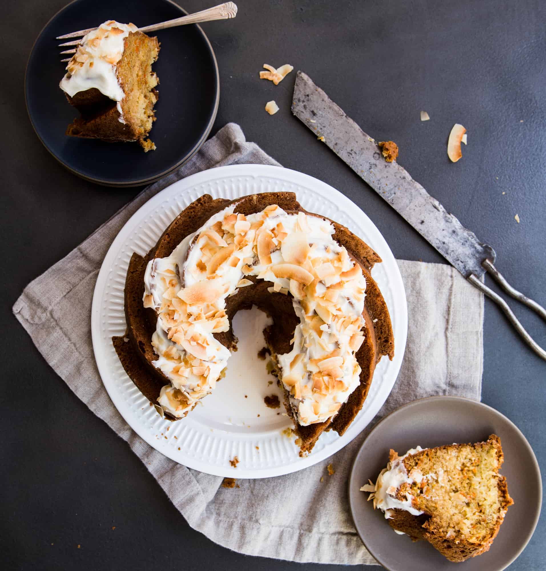 Dark background. carrot Bundt cake on a white plate with two slices removed. Slices sitting on a dark plate with a fork and a tan plate with no fork. Slicing knife on the side 