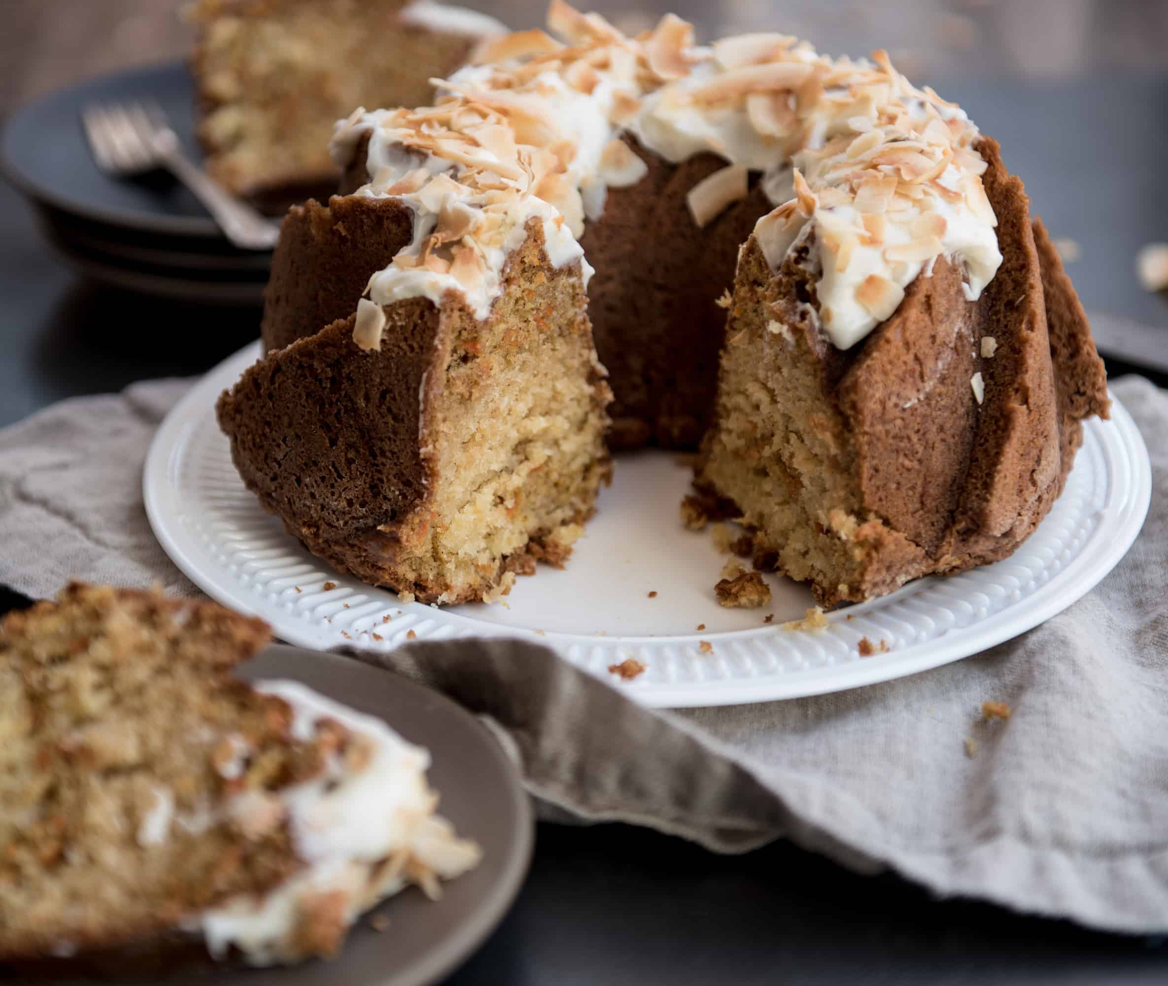 Close up of a sliced carrot bundt cake that reveals the swirl and the crumb in the center. Cake is on a white serving plate sitting on a tan linen napkin on a dark colored table. 