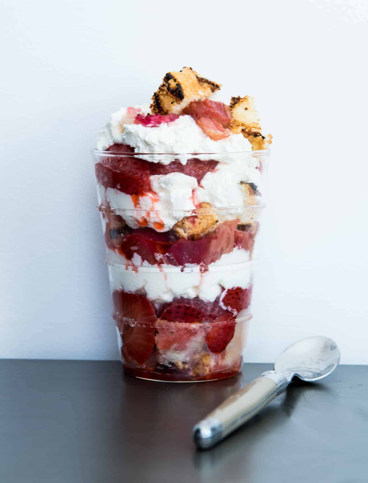 A clear glass containing a grilled rhubarb and strawberry parfait. Layers of grilled angel food cake, whipped cream, grilled strawberries and grilled rhubarb