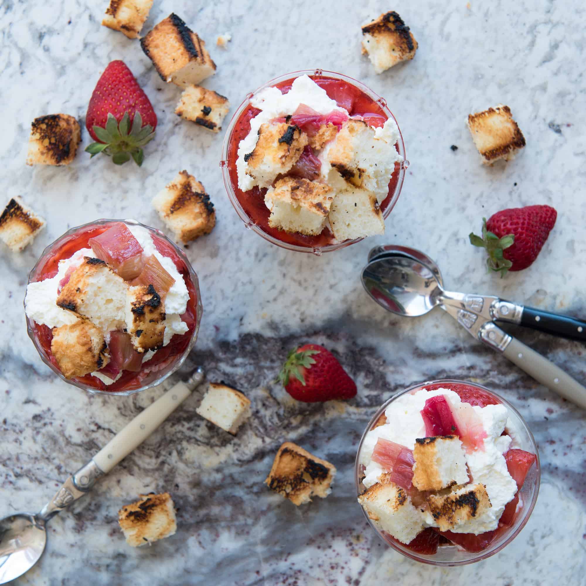 overhead of three finished grilled rhubarb and strawberry parfaits. Whole strawberries, sliced grilled angel food cake and spoons scattered around. 