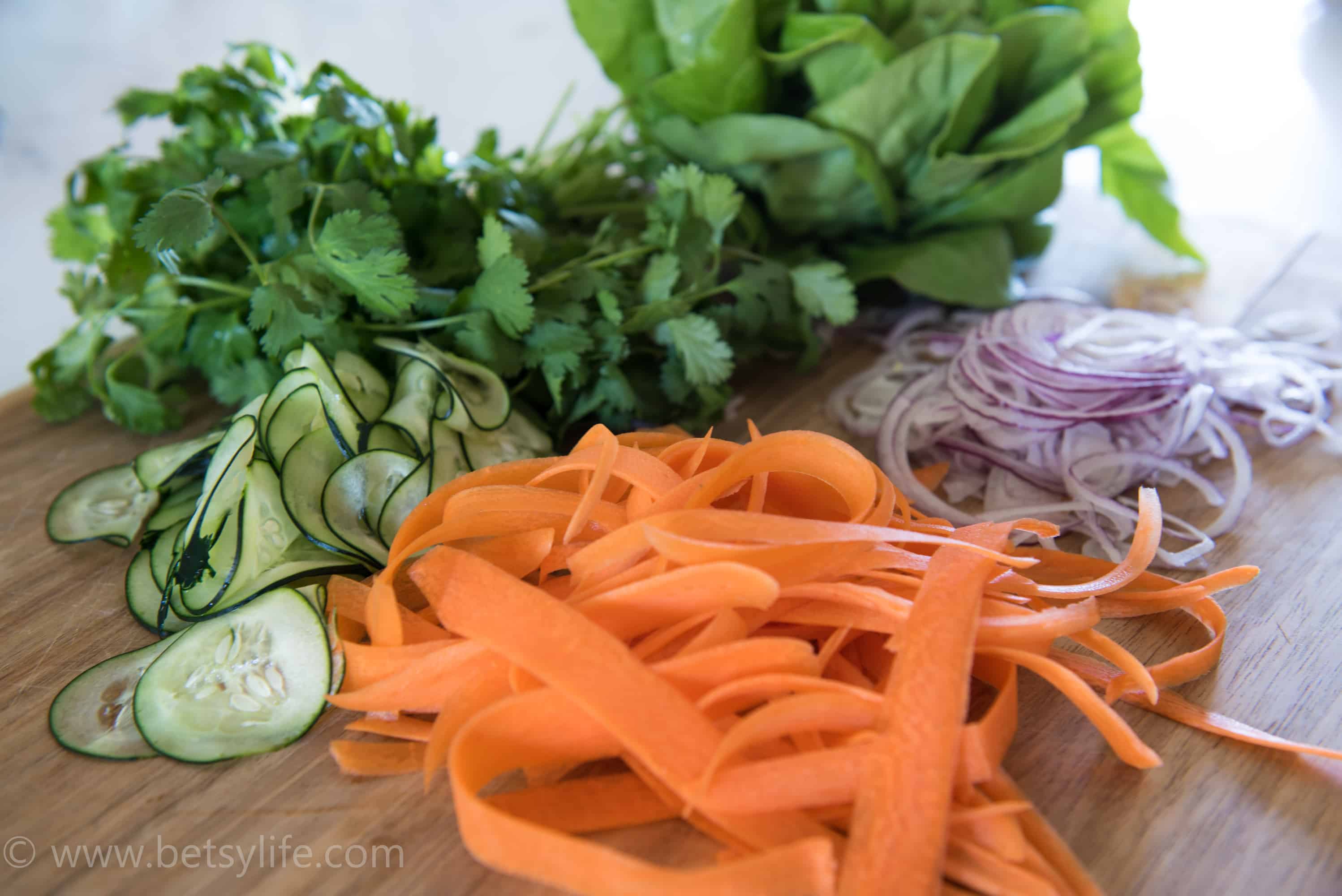 wooden cutting board piled with thinly shredded carrots, cucumber slices, thin slices of red onion, a bunch of cilantro and a head of butter lettuce 