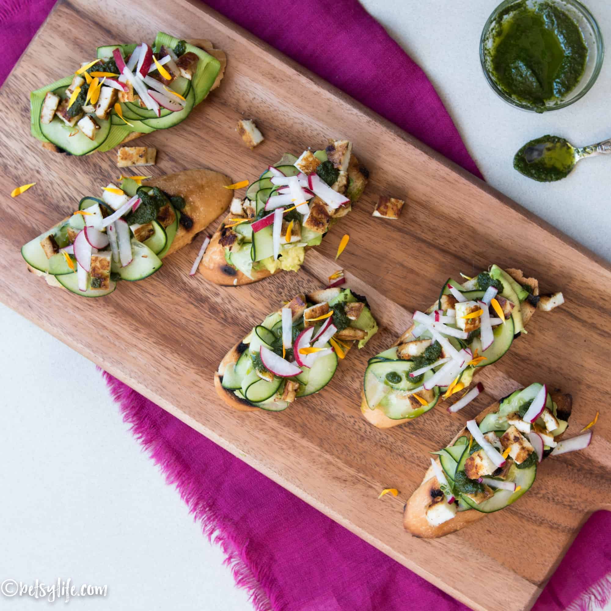 overhead of 6 avocado toasts on a wooden board topped with grilled halloumi cheese chunks, cucumber, radish and edible flowers. Jar of herb oil with a spoon on the top right corner of the photo 