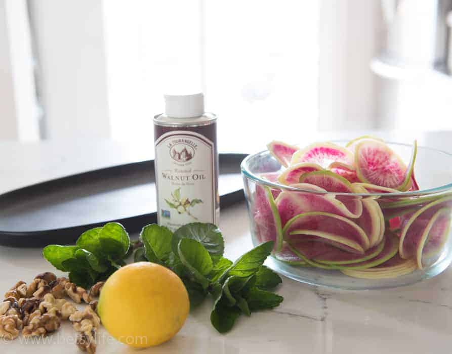 white counter top with oval black plate, pile of walnuts, bunch of mint, whole lemon, bottle of walnut oil and a glass bowl filled with thinly sliced watermelon radishes 