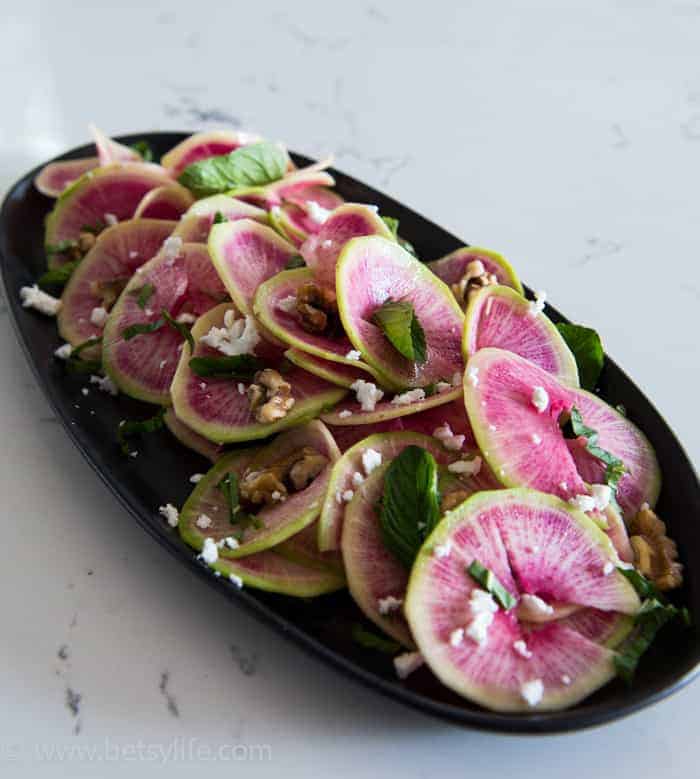 thinly sliced watermelon radish salad on a oval black plate. Garnishes with mint leaves and feta cheese crumbles 