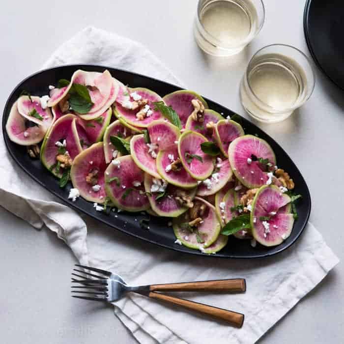 Overhead photo of thinly sliced watermelon radish salad on an oval black plate on top of a natural linen napkin. Light colored background. Two glasses of white wine in the upper right corner and two forks with wooden handles in the lower left corner 