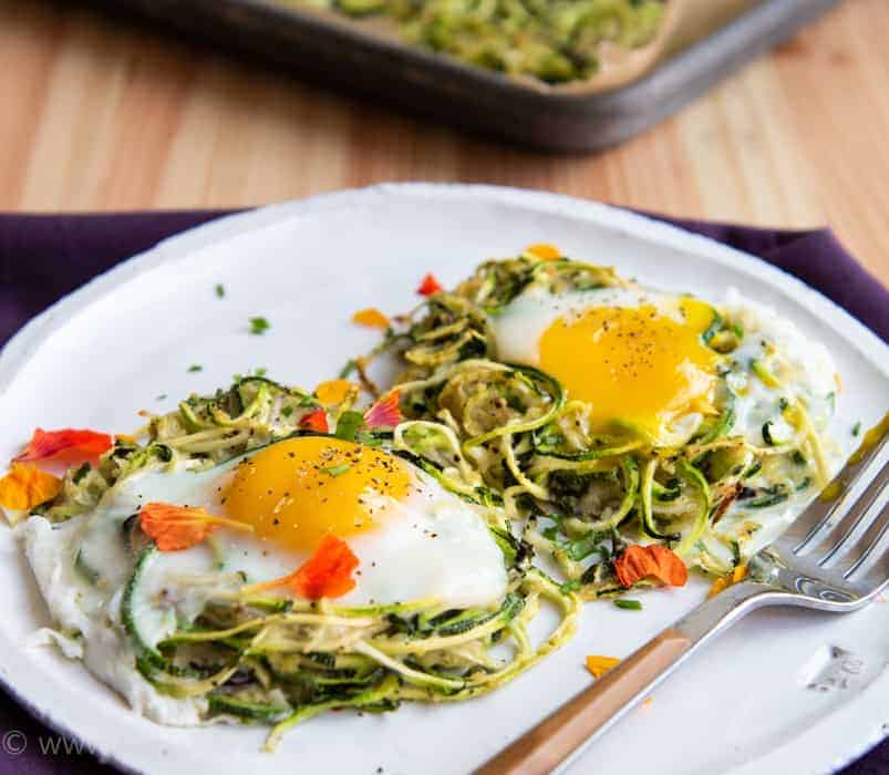 Two Spiralized zucchini nests on a white plate with runny yolk eggs on top. Fork to the right and a purple napkin underneath
