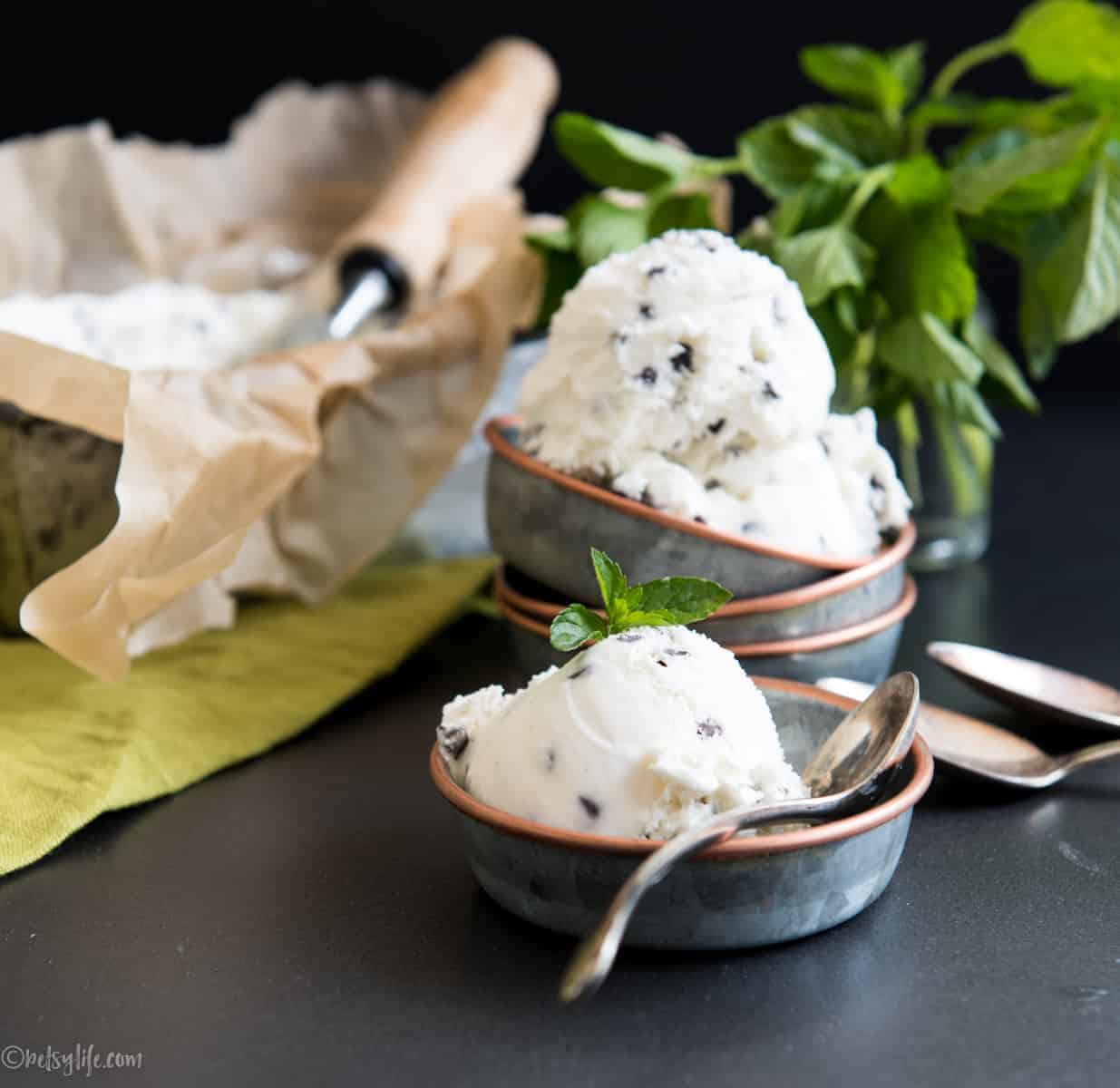 two metal bowls of mint chip ice cream on a dark surface with fresh mint in the background