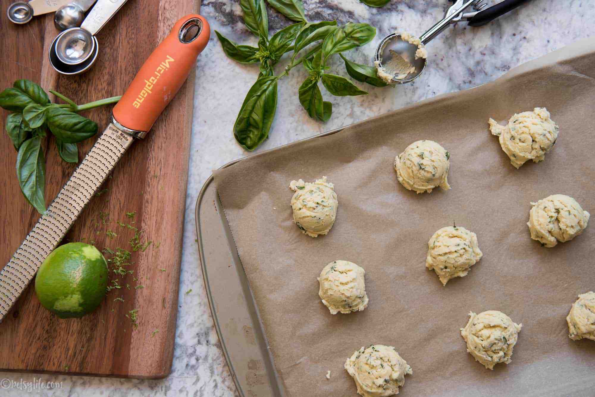 Tray of unbaked balls of basil lime sugar cookie dough next to a cutting board with lime and basil
