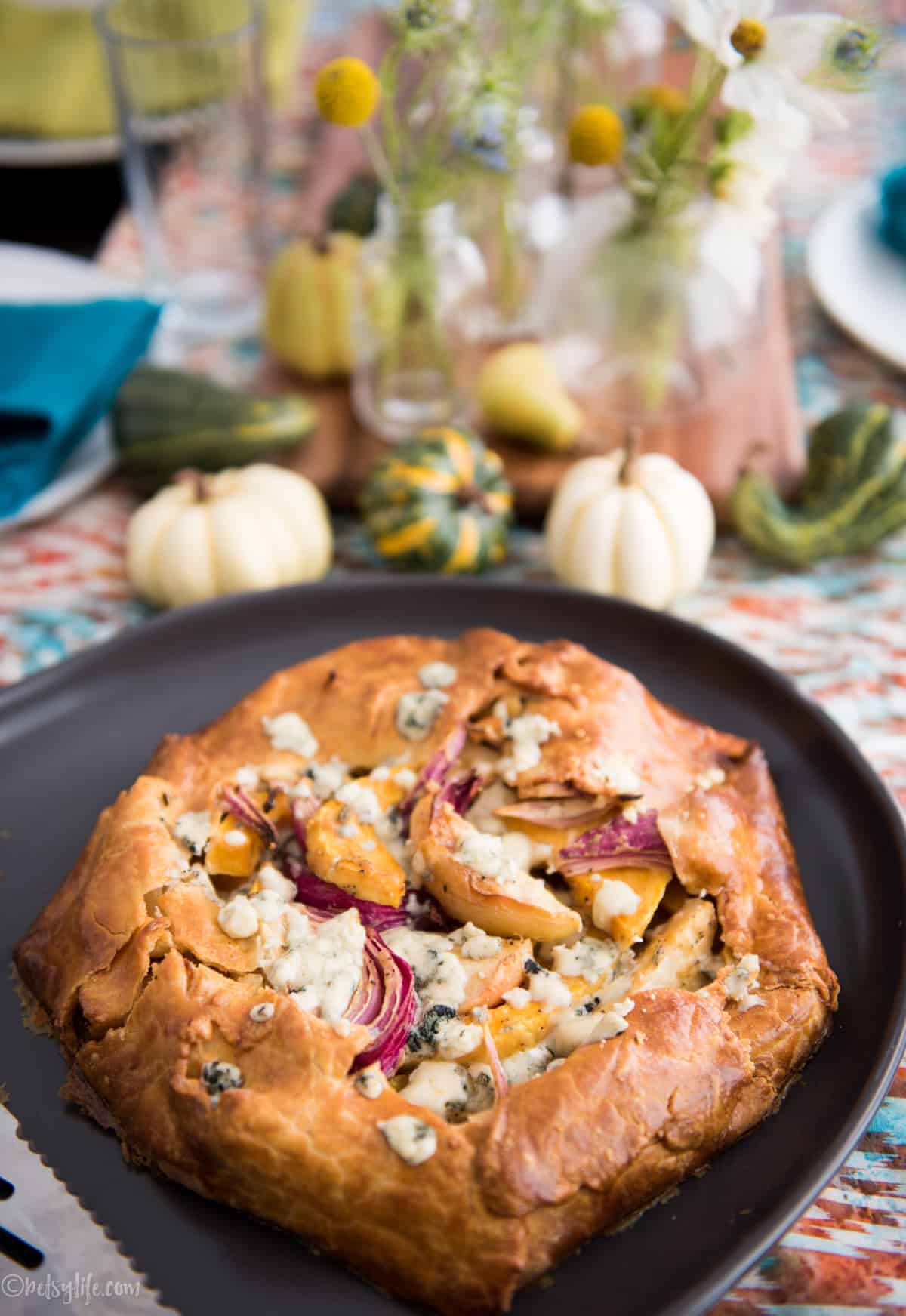 savory apple galette on a brown serving plate with flowers and gourds in the background