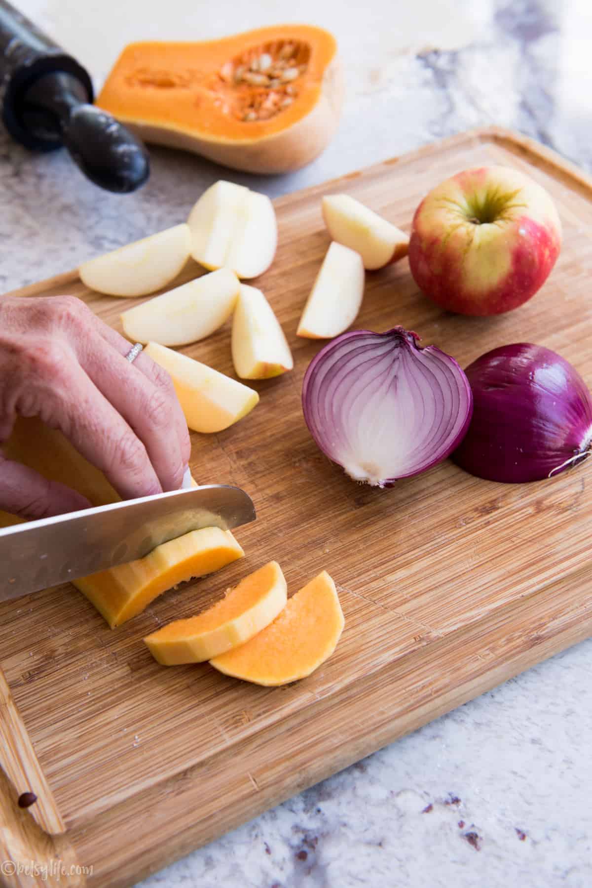 cutting board with butternut squash being sliced. Onions and apples on the side. 