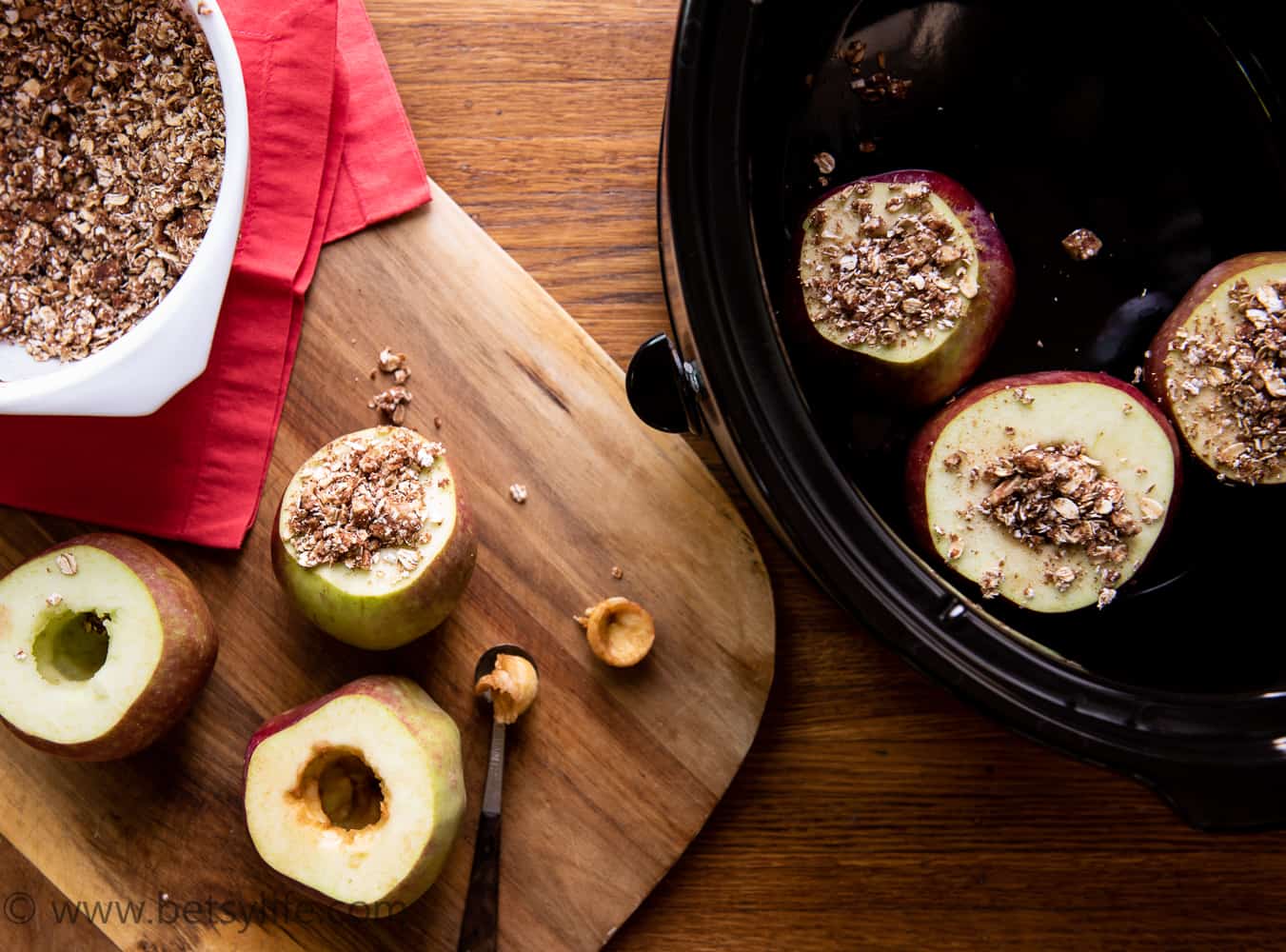 Baked apple preparation. Bowl of oat mixture, cored apples, several stuffed with mixture on a cutting board next to a crock pot