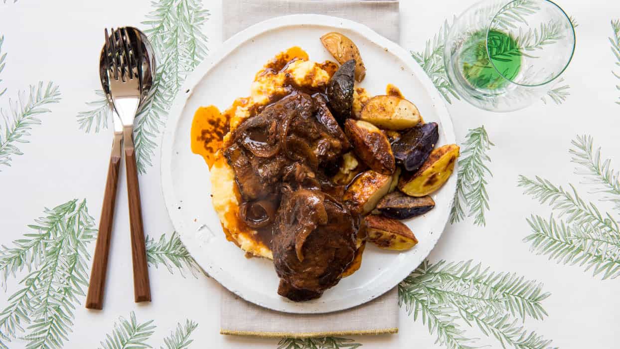 White plate with polenta, roasted potatoes and beef short ribs on a white background printed with green pine boughs 