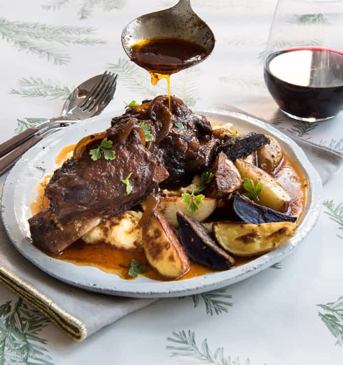 Plate of beef short ribs, roasted potatoes and polenta with a spoon drizzling sauce over the top 