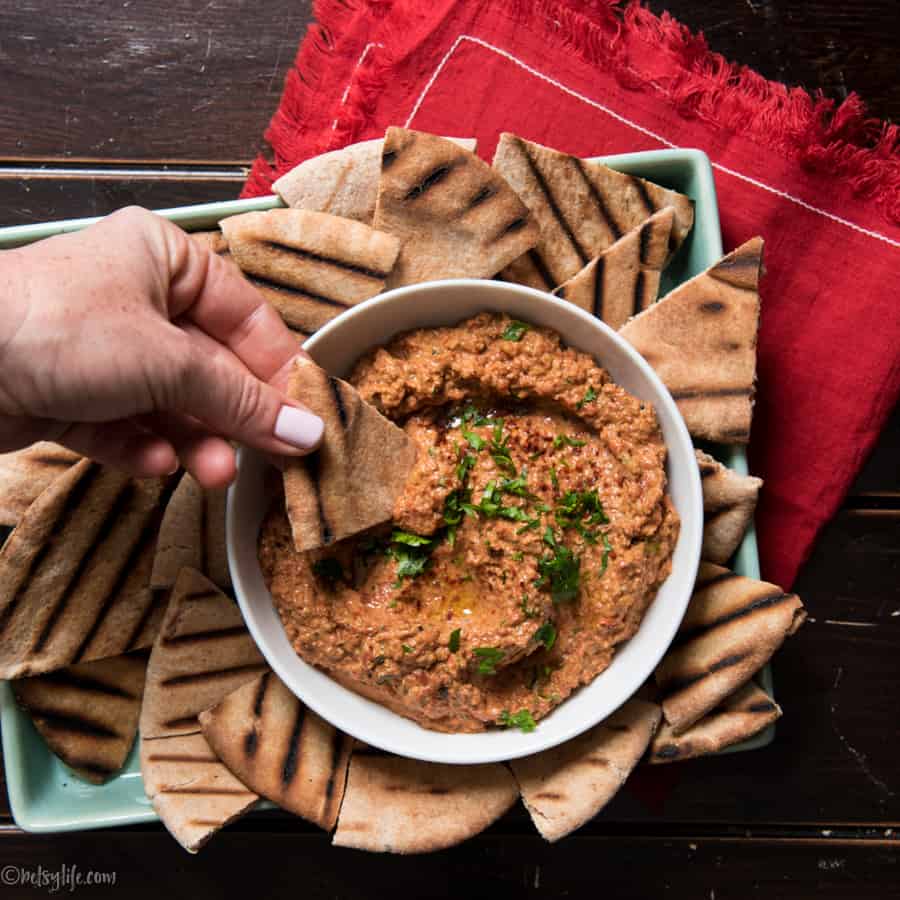 hand dipping a pita into a bowl of muhammara roasted red pepper and walnut dip on a platter of grilled pita bread cut into triangles