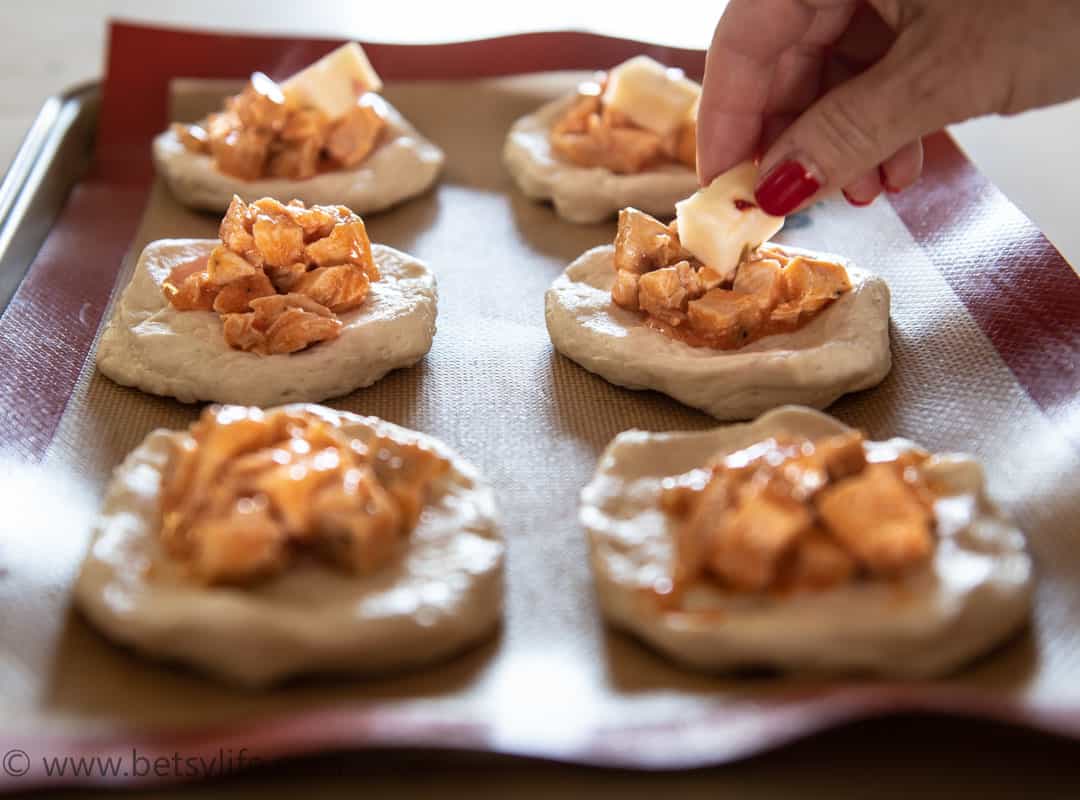 Tray of biscuit dough rounds topped with buffalo chicken. Hand placing cube of cheese on top. 