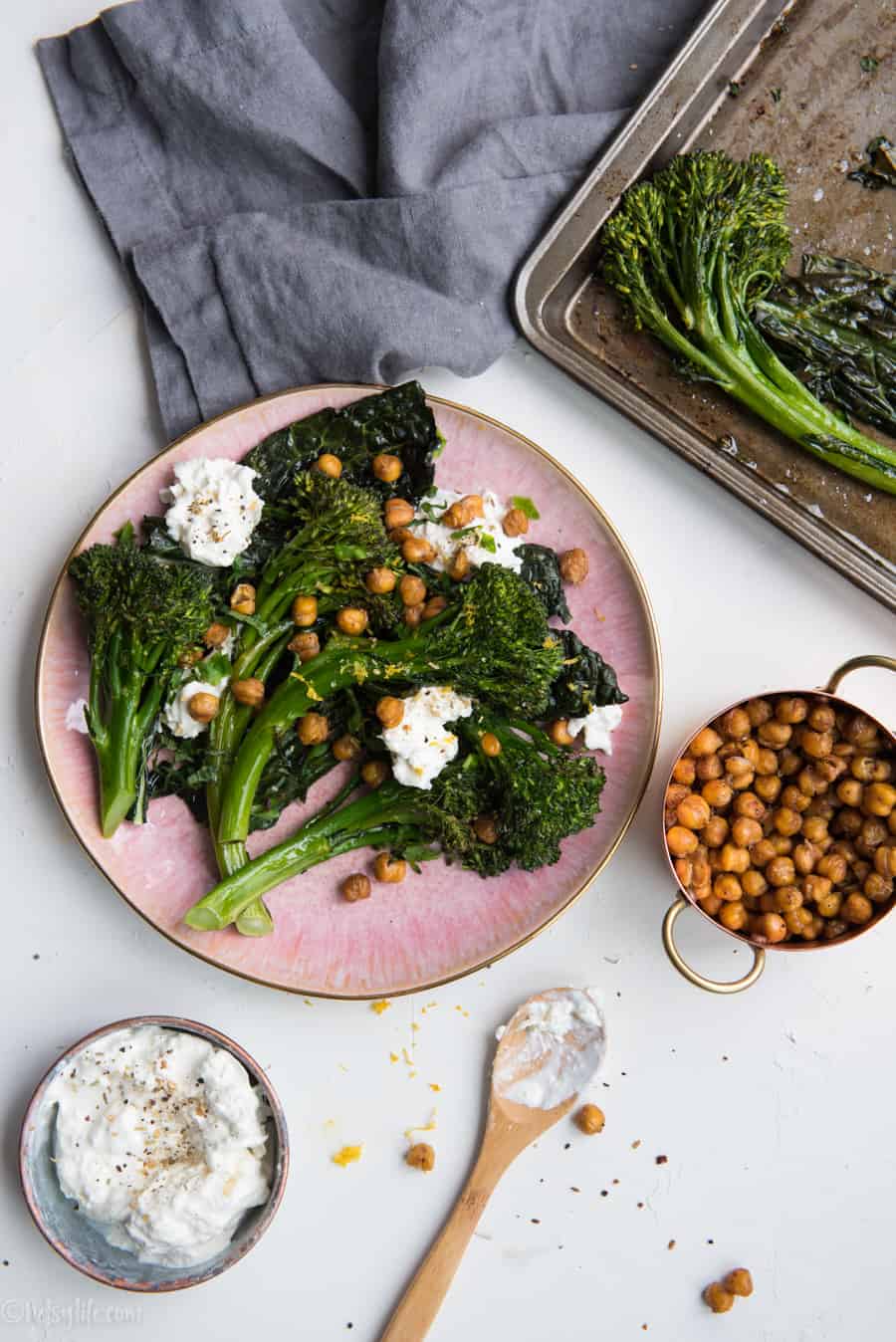 Roasted Broccolini with Crispy Kale, Chickpeas and Burrata on a pink plate next to a roasting pan, a gray linen, a measuring cup filled with chickpeas and a metal dish filled with cheese next to a wooden spoon