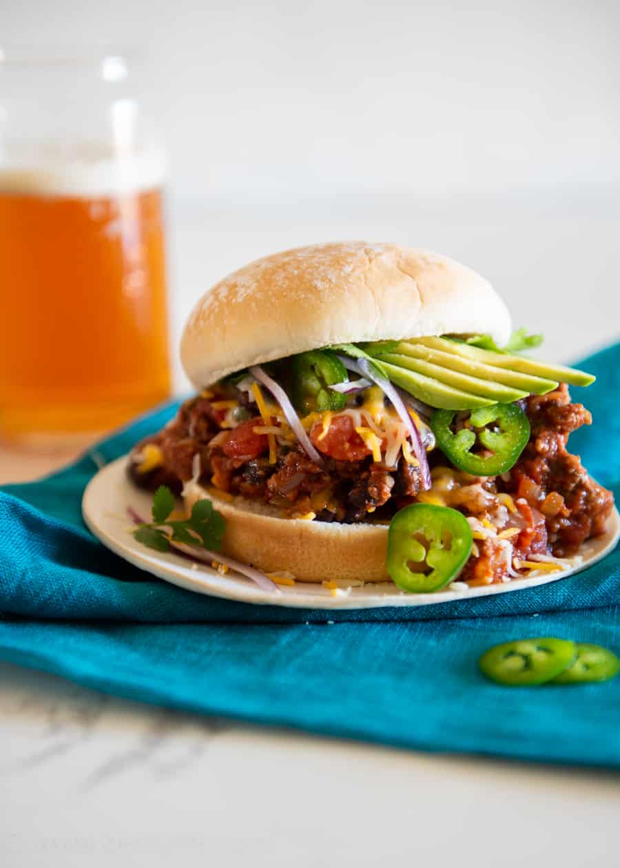 Southwestern style spicy sloppy joe piled high on a bun with melted cheese, sliced avocados, jalapenos and red onions next to a glass of beer