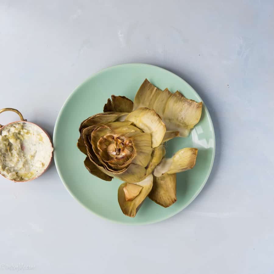 roasted artichoke on a green plate next to a small dish of cheese dip 