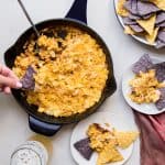 skillet of roasted corn dip next to plates with dip and tortilla chips. Hand dipping into skillet with a blue tortilla chip