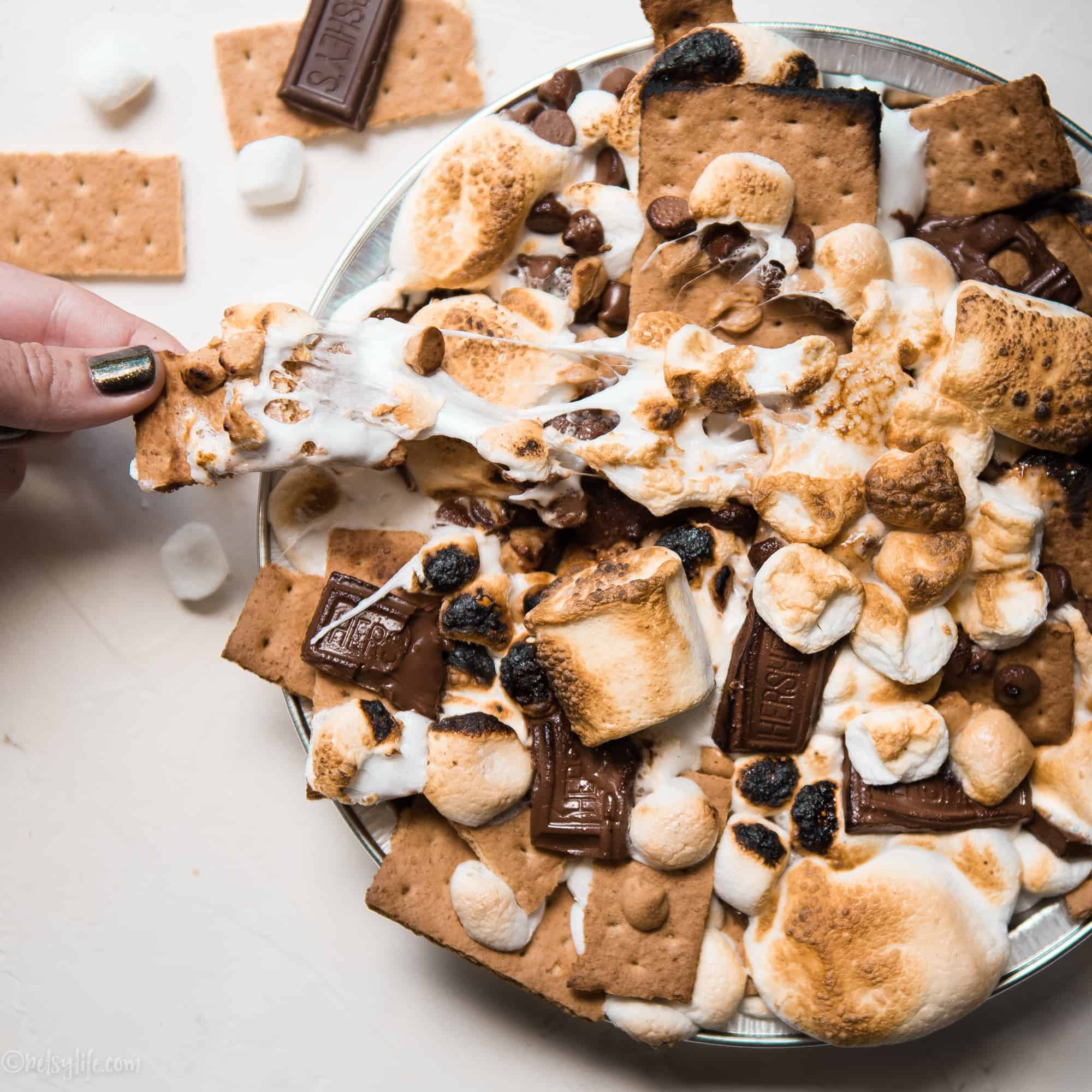 Camping recipe. Hand pulling a gooey marshmallow covered graham from a round dish of s'mores nachos