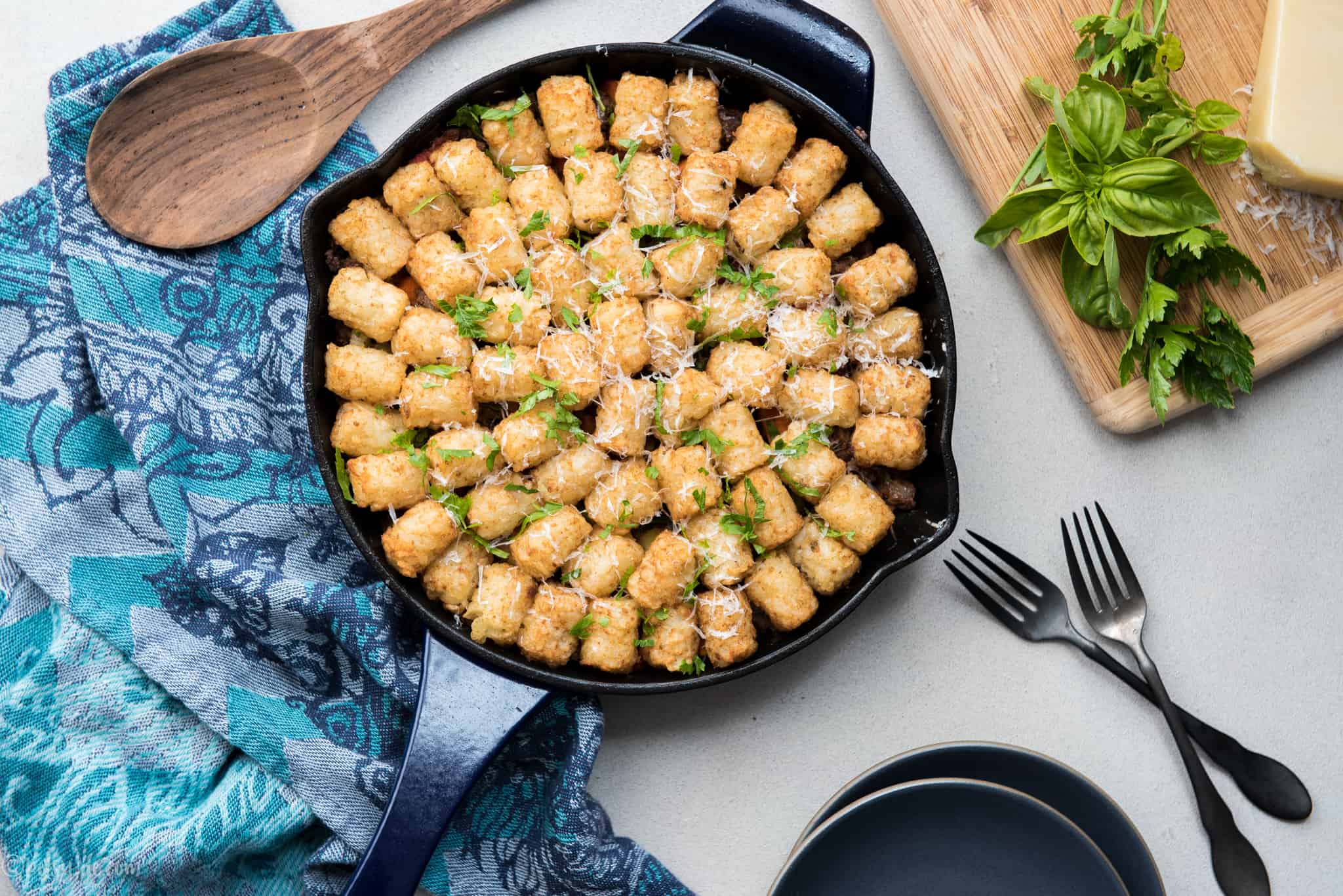 Skillet casserole topped with tater tots next to plates, forks and a cutting board with cheese and herbs
