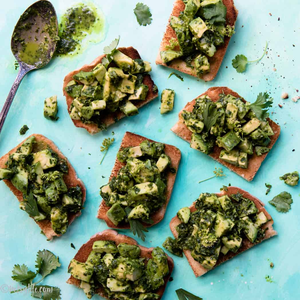 Avocado bruschetta with chimichurri and herbs on a blue background with a spoonful of chimichurri on the side