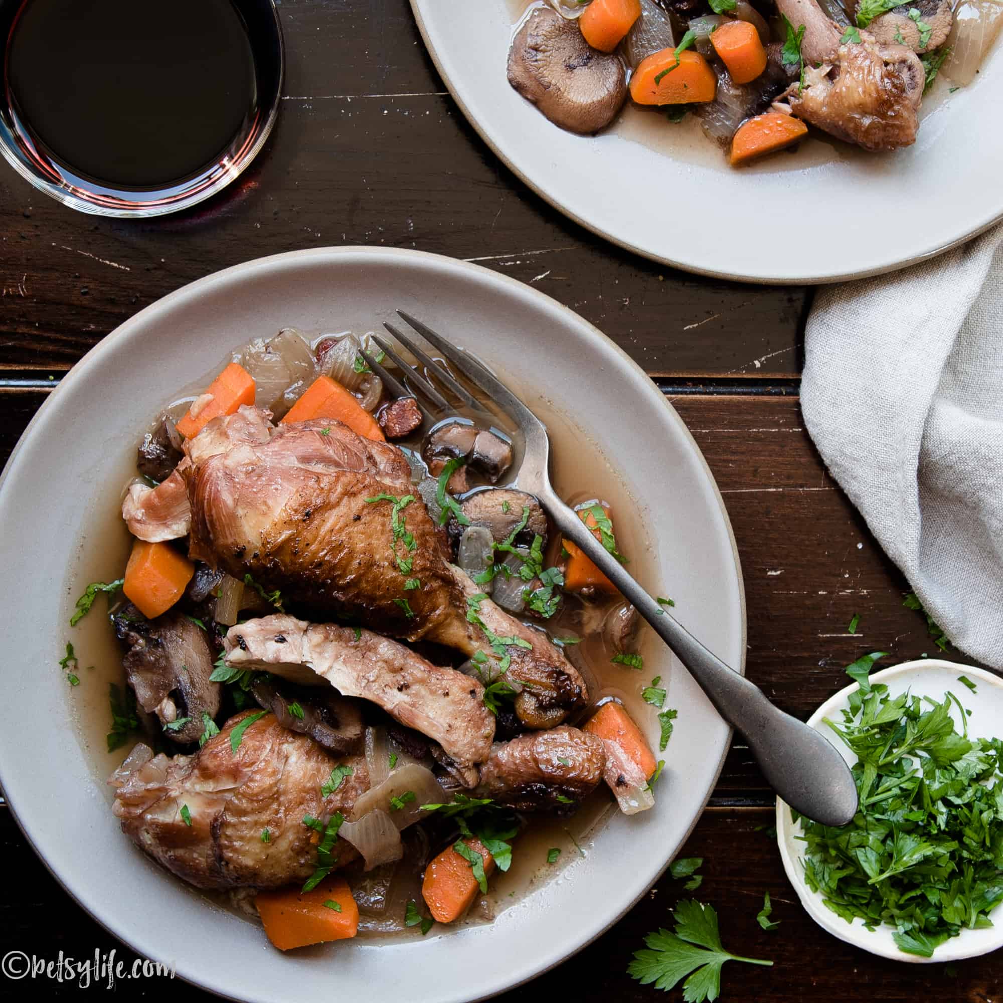Plate with Coq au vin and and veggies next to a glass of wine and fresh herbs