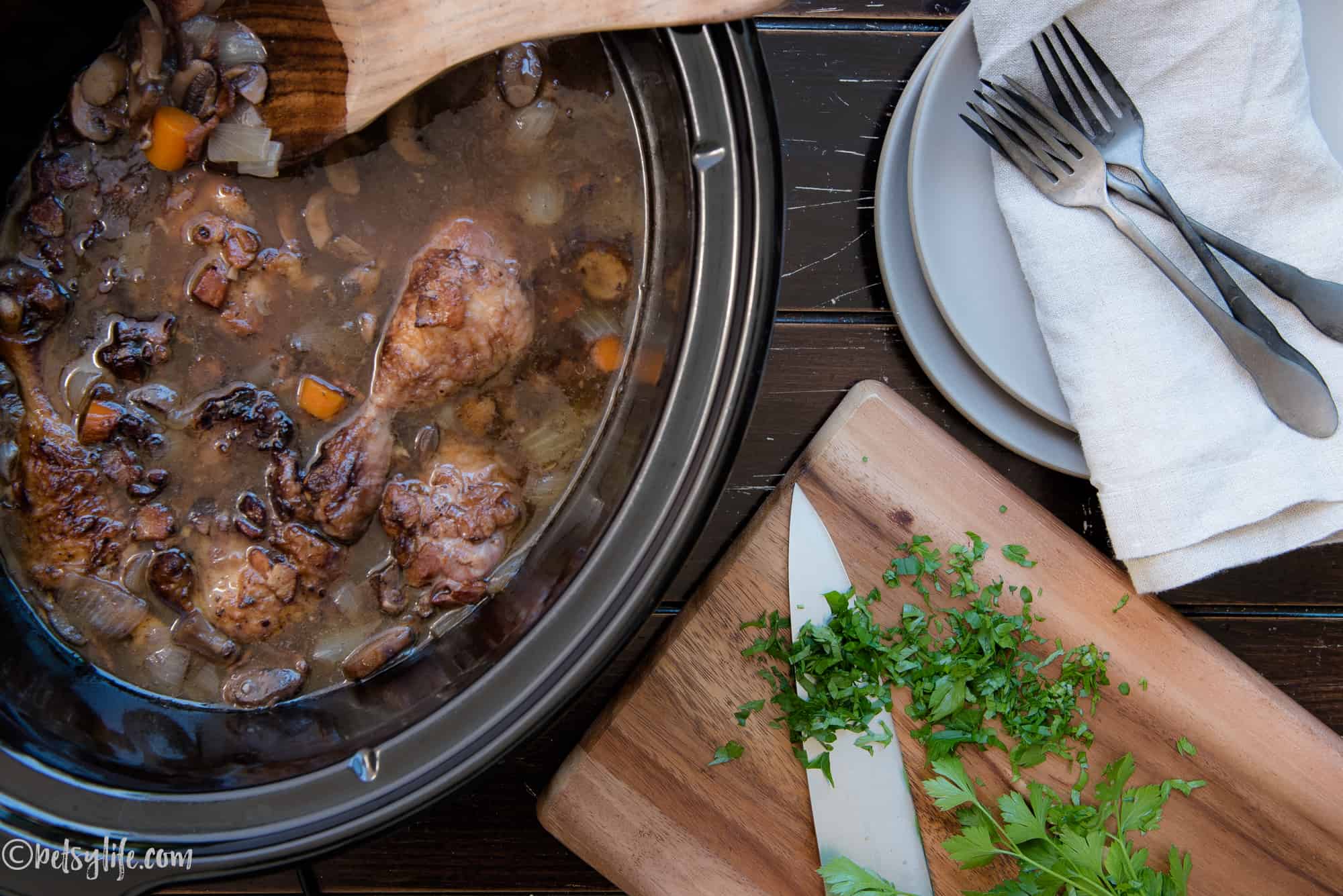 Crock pot filled with coq au vin next to stack of plates and cutting board with herbs 