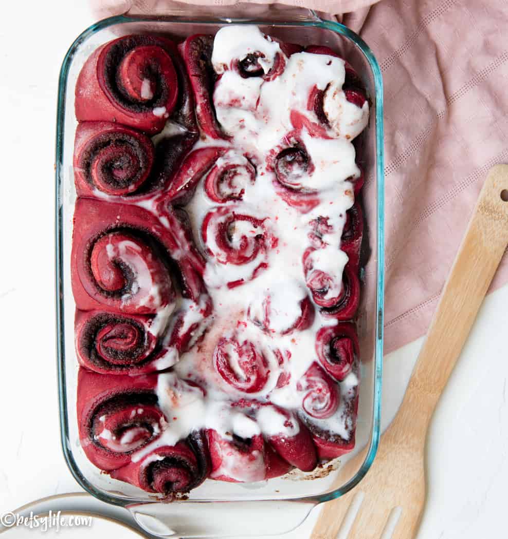 Baking dish of red velvet cinnamon rolls that is half covered in frosting 