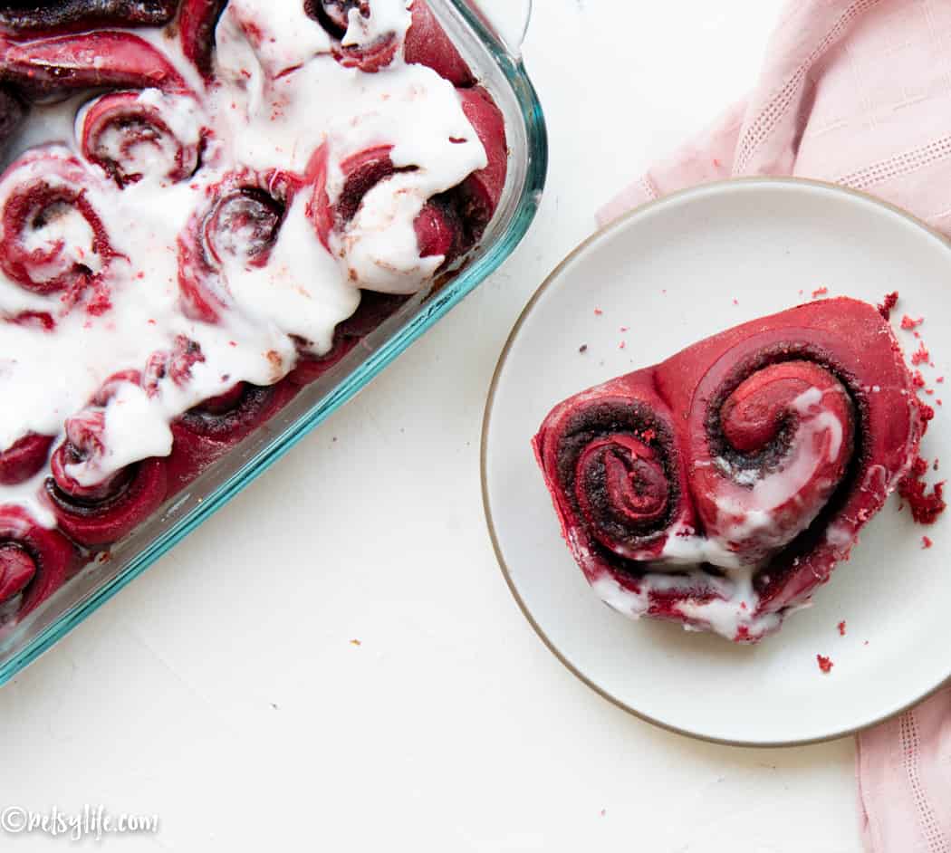 Heart shaped red velvet cinnamon roll on a white plate next to a baking dish of more cinnamon rolls 