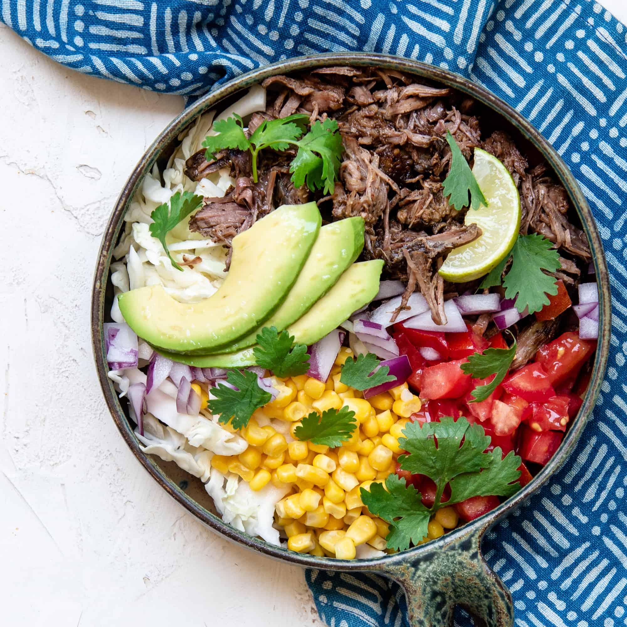 Burrito bowl with spicy beef barbacoa, avocado, tomatoes, lime, cilantro, cabbage and corn next to a blue pattern napkin
